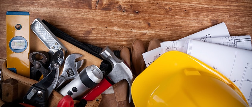 A collection of tools including a hammer, pliers, level, flashlight, gloves, and a yellow hard hat on a wooden surface.