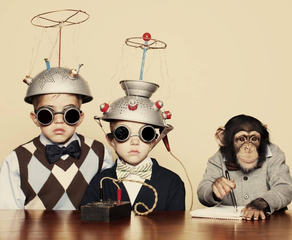Two children in quirky outfits with colanders on their heads and goggles, alongside a dressed monkey writing in a notebook.